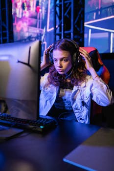 Focused young woman gaming at a computer setup with headsets, indoors.